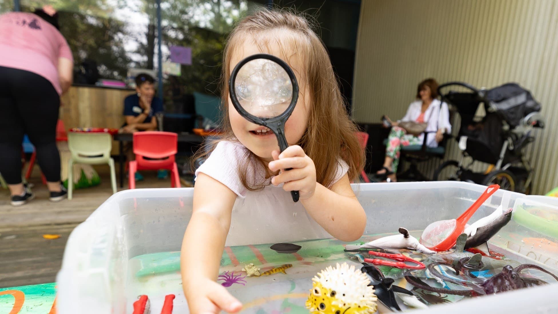 Toddler girl looks through magnifying glass over water table at Discovery Garden