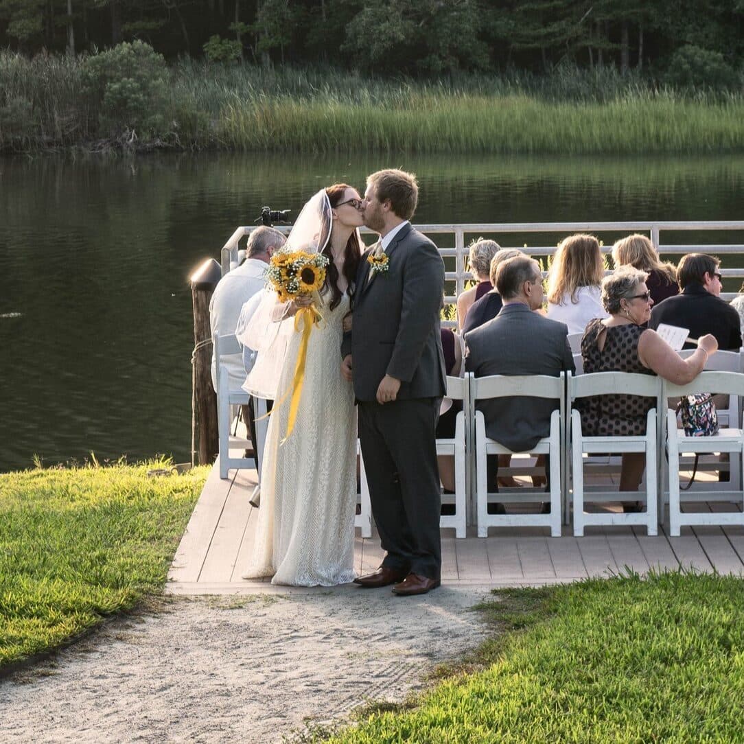 A bride and groom kiss behind their wedding guests outdoors in front of Owls Creek